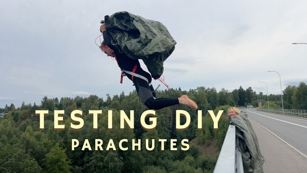 A person wearing a black wetsuit is jumping off a bridge while holding a DIY parachute made from a tarp. Another person looks on from the bridge, partially wrapped in a similar tarp. The text “TESTING DIY PARACHUTES” is overlaid on the image. The backdrop shows greenery and trees under a cloudy sky.