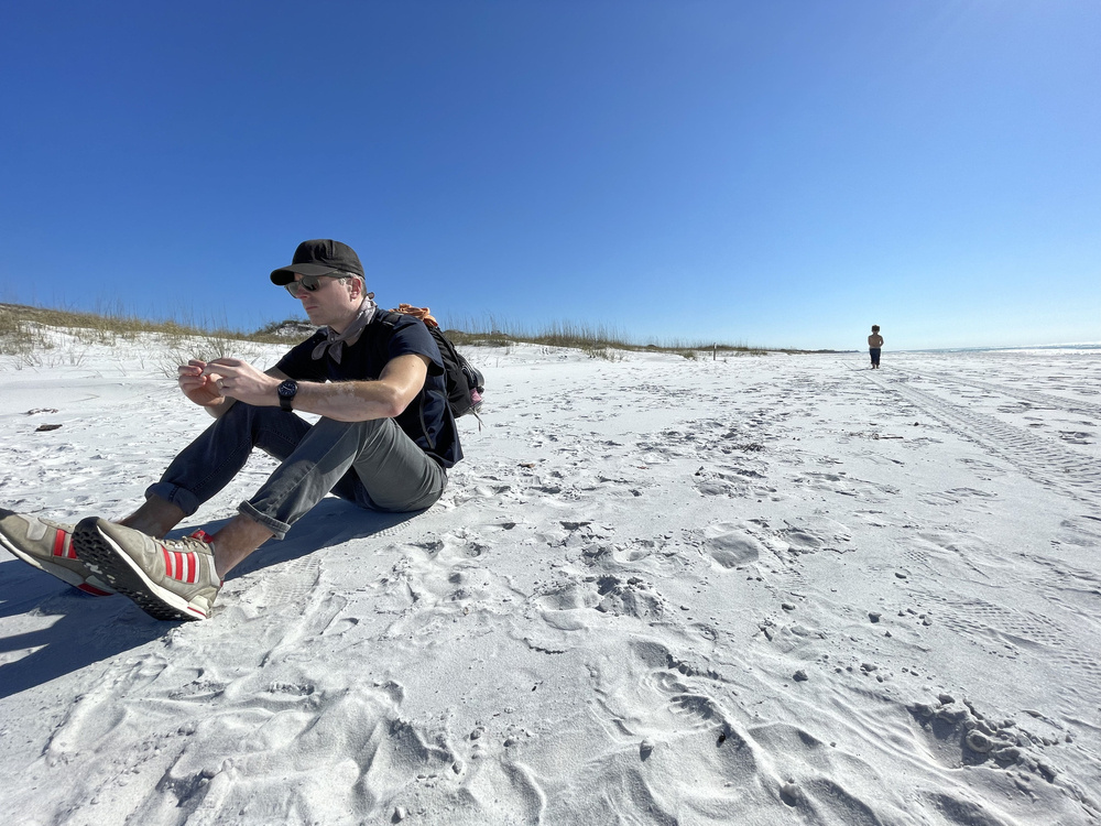 Man on a beach with withe sand wearing a black hat, sunglasses, t-shirt, and jeans.  Small child in the background.