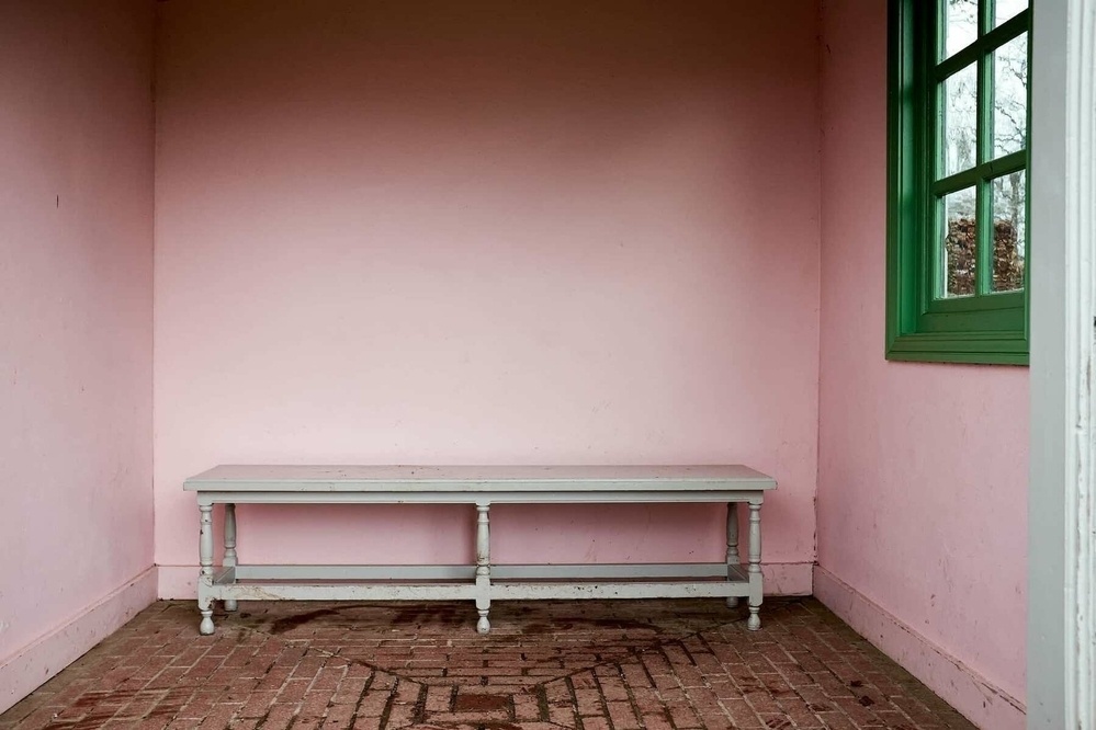 A tranquil outdoor room with one side open to the outdoors. A simple wooden bench is placed in front of the soft pink rear wall, and there is a window with a green frame to the right of the photo.