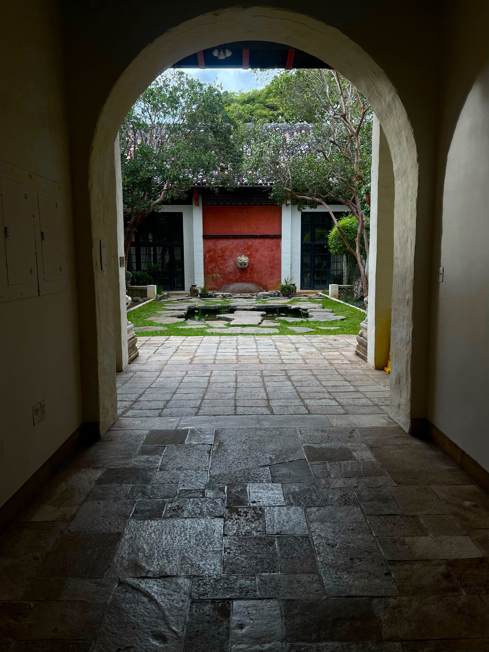 A courtyard framed by an arched doorway, featuring a red door, greenery, a pond and a stone path.