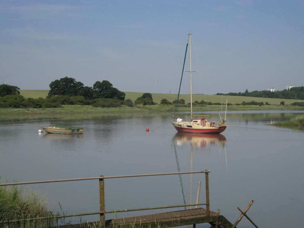 A rowing boat and small sail boat moored in a river estuary. In the foreground is a small rusty walkway. In the background are rolling green meadows and buildings just poking over the trees.