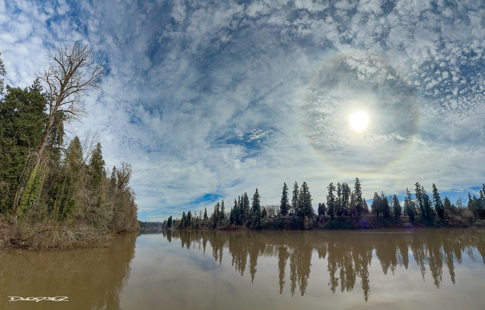 A scenic river reflects a forested shoreline and a cloudy sky with a faint sun halo.