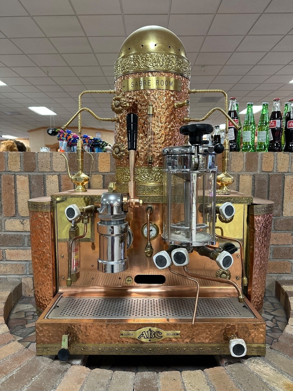 An ornate espresso machine made of copper and brass with decorative elements, labeled “Sabre Room,” on top of a brick counter with bottled drinks visible in the background.