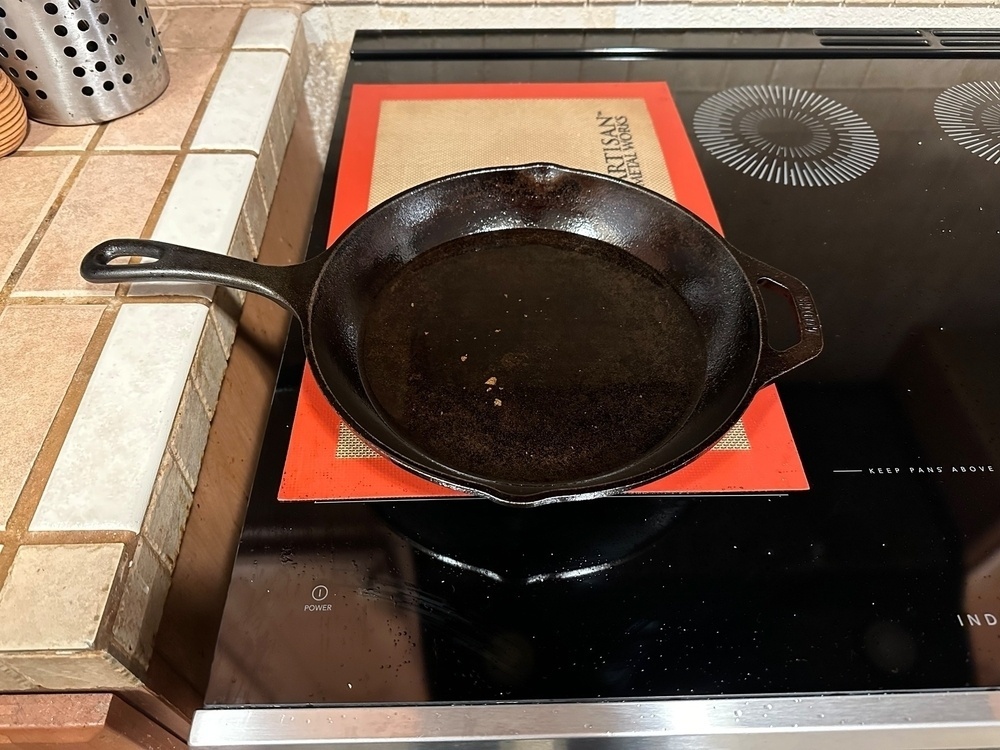 A cast iron skillet on an induction stovetop with a silicone mat underneath and a perforated stainless steel utensil holder at the side.