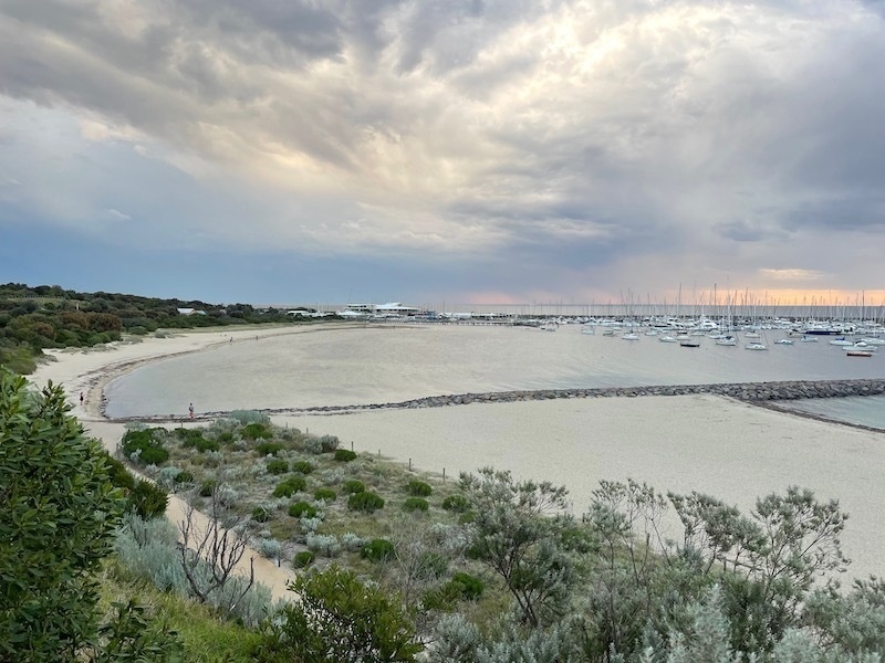 a view overlooking a beach, with light grey-blue water and cloudy skies with some peach-coloured sky on the horizon