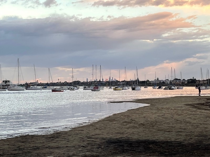 a view on the beach; a bunch of boats are floating in the water, the sky has got more pink, and in the distance sunlight is reflecting off towers in the distant city