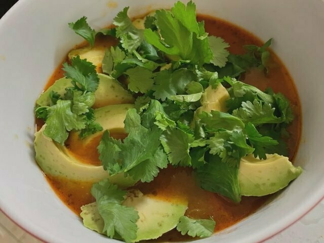close-up of a bowl of tomato-y soup, garnished generously with sliced avocado and fresh coriander