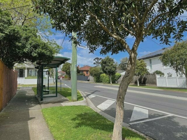 a suburban street. a sheltered bus stop with a bench is prominently in view. beyond it lies a T intersection, with detached houses all around. leafy trees frame the shot.