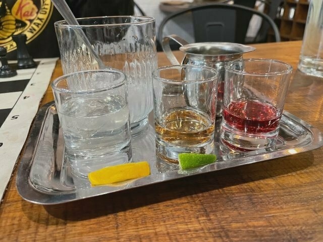 A tasting tray with three varieties of gin – one clear-coloured, one brown, one red – with a glass of ice and a little pouring jug of tonic water.