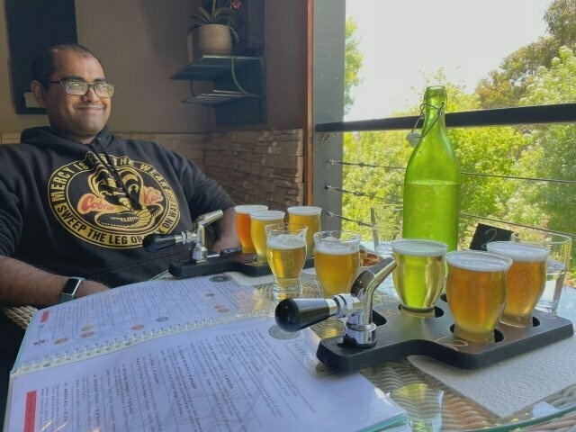 An Indian guy smiling tight-lippedly at the camera, sitting on a shaded deck overlooking abundant greenery, with a table in front of him with two pallets each with four different beers