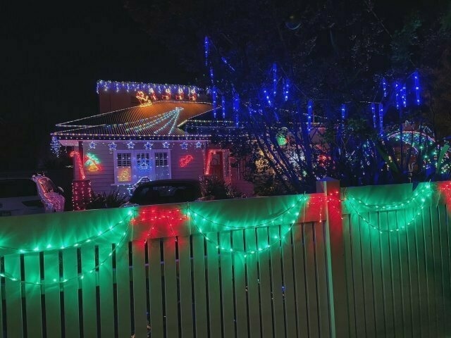 A house with lots of multicoloured lights all over it; there are also blue lights throughout a tree in front of the house and green and red lights along the front fence (the red lights are bow-shaped)