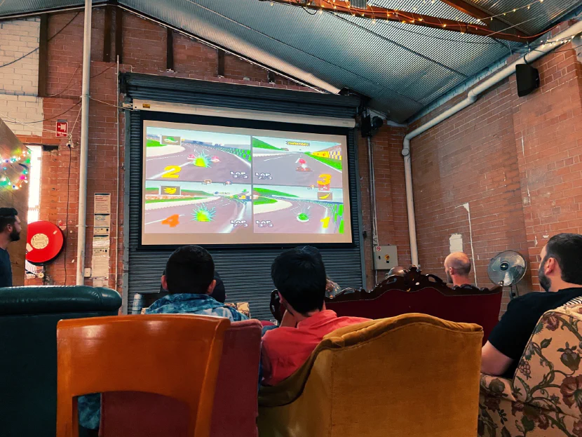 a game of Mario Kart 64 is being projected on a screen inside a pub with brick walls. at the bottom of frame are the backs of a few seated guys' heads