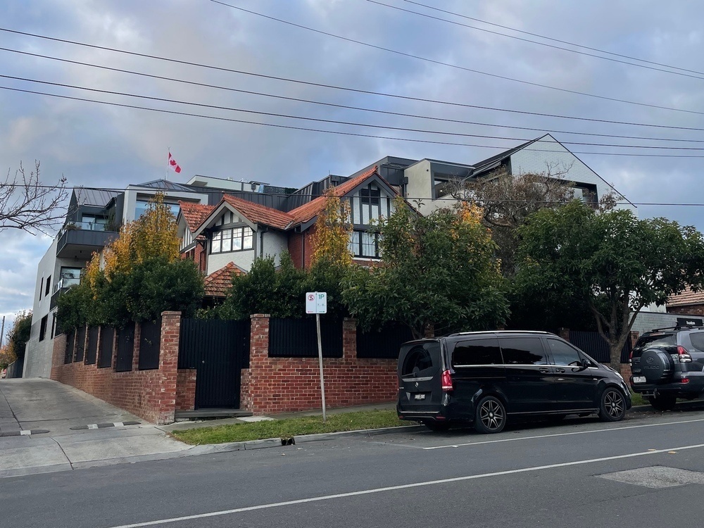 Looking across the road at a building, which looks like a two-storey house at the front and a modern white/dark grey apartment block in the rear. Above one of the rear apartments is a Canadian flag.