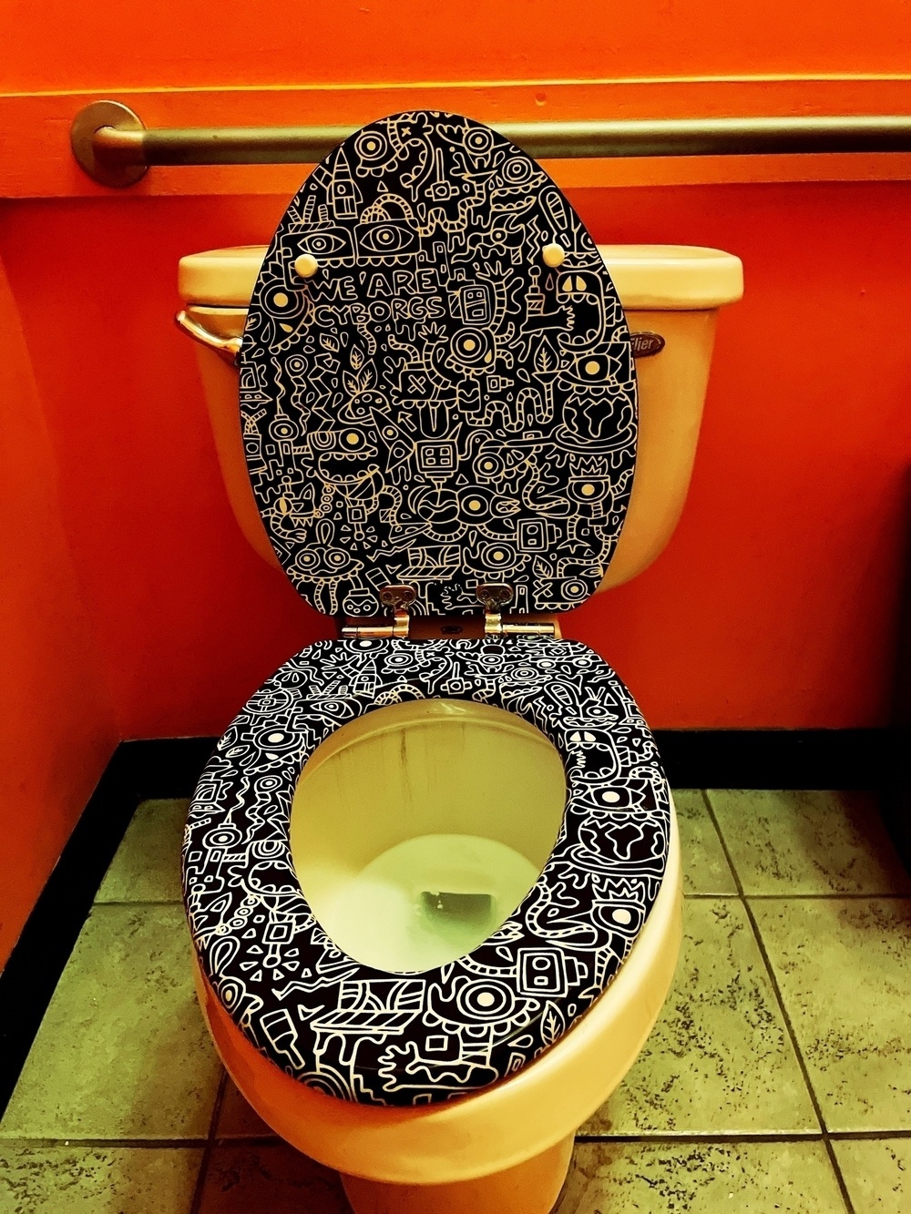 A toilet with a seat and lid covered in intricate black and white doodles is set against a vibrant orange wall.