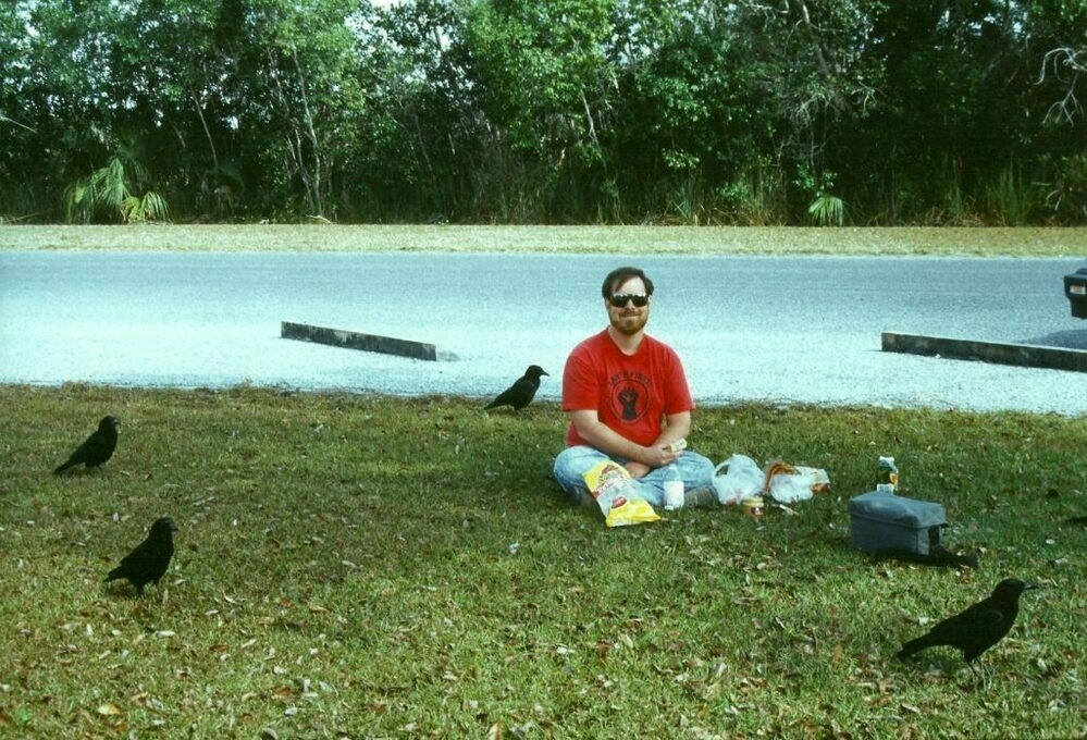 Me in a red shirt sitting on the ground with a picnic lunch, surrounded by crows