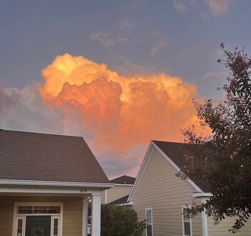 against a blue sky, a billowing cloud lit fire-orange by the sunset, over two simple suburban houses.