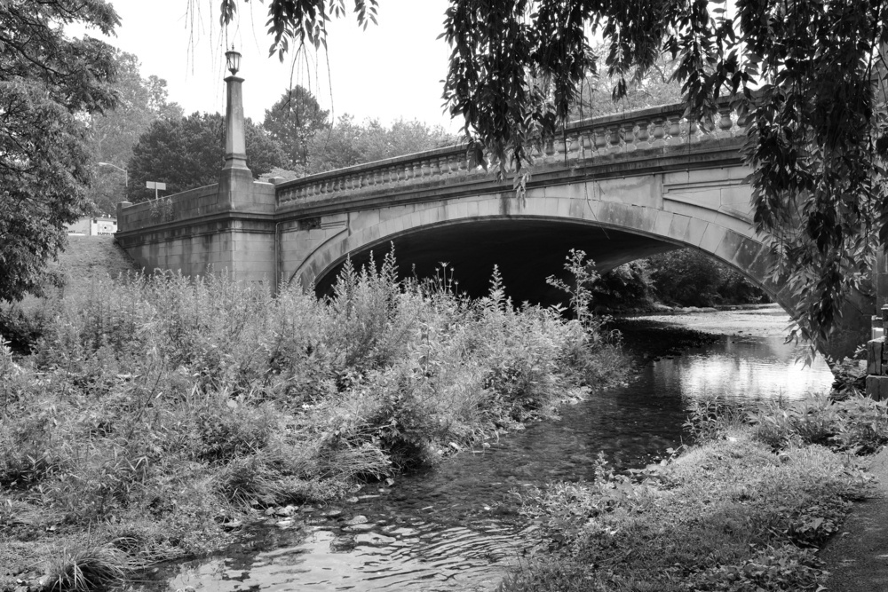 Black and white photo of a classically styled concrete bridge crossing a large stream with foliage.
