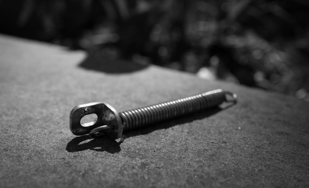 Black and white photo of a metal spring laying on a stone wall with a bracket on the left end.