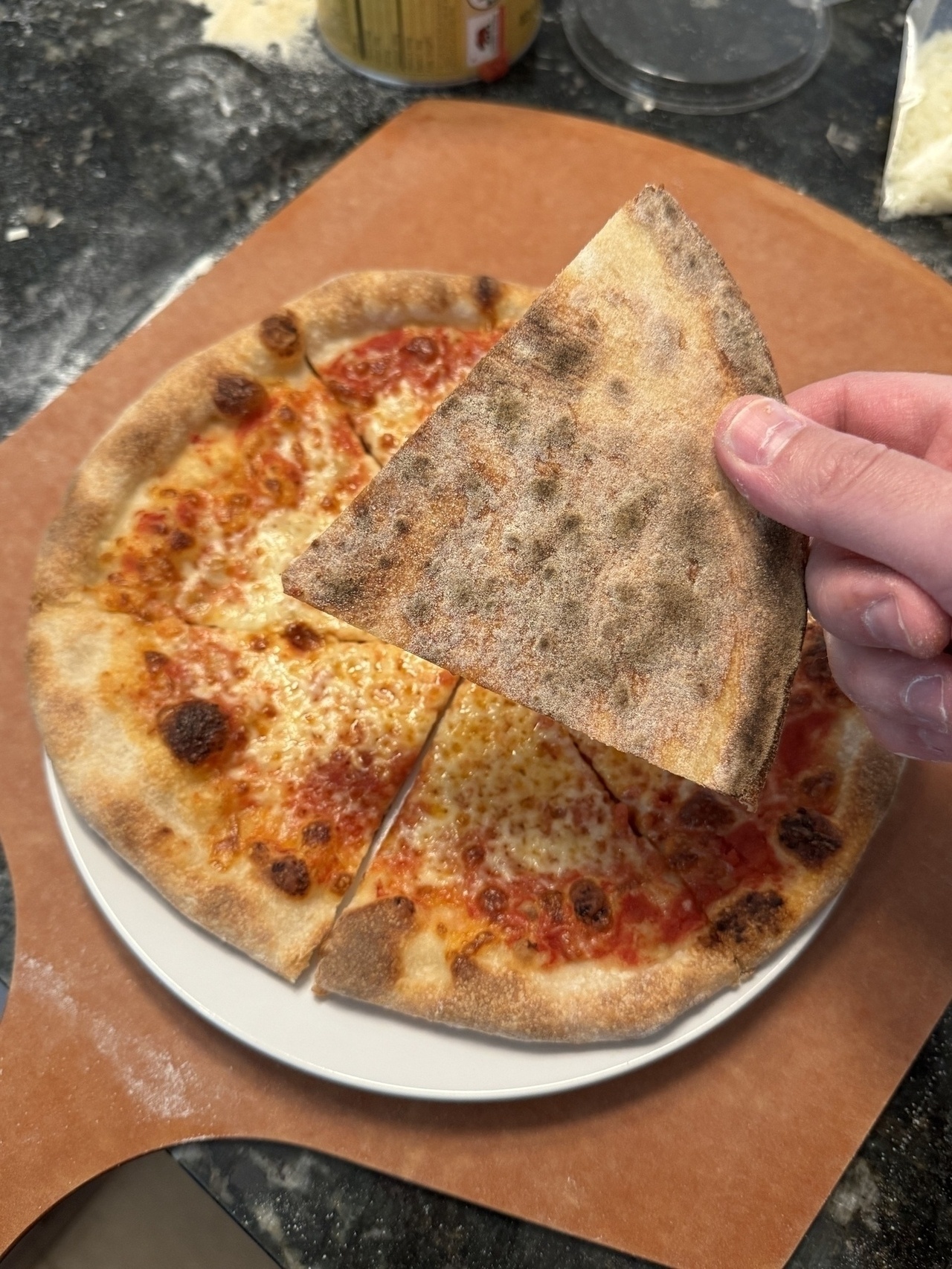 A hand holds a slice of pizza with a crispy crust above a plate of the remaining pizza.
