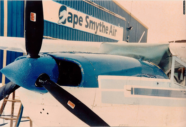 A propeller airplane is parked in front of a building with signage that reads Cape Smythe Air.