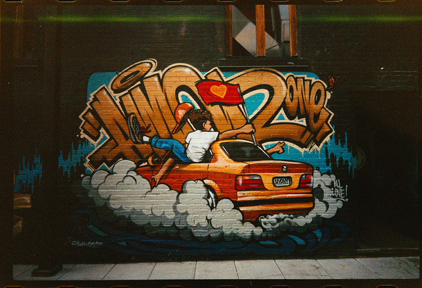 A vibrant mural depicts a person joyfully riding on the back of an orange car with smoke billowing around it, set against stylized graffiti text.