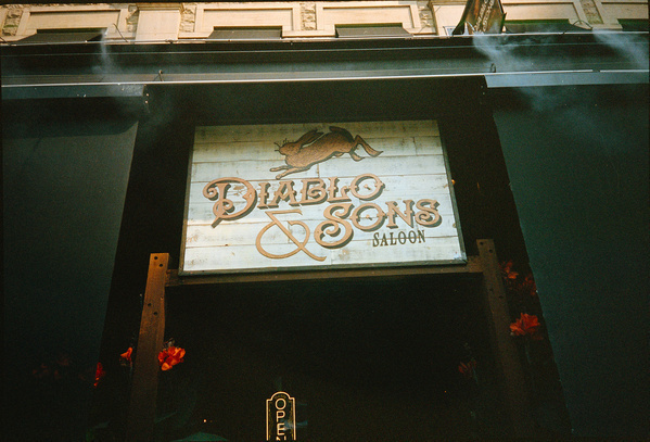 A wooden sign with the text Diablo & Sons Saloon featuring a leaping wild boar is mounted above a dark entrance with an Open sign below.