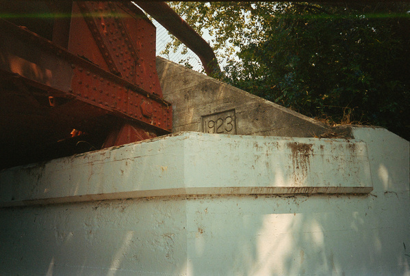 A concrete structure with the year 1923 engraved on it supports a rusty red metal bridge beam surrounded by trees.