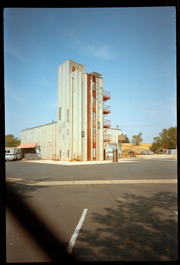 A tall, narrow building with exterior staircases is situated in a mostly empty paved lot with other industrial structures and some vehicles in the background.