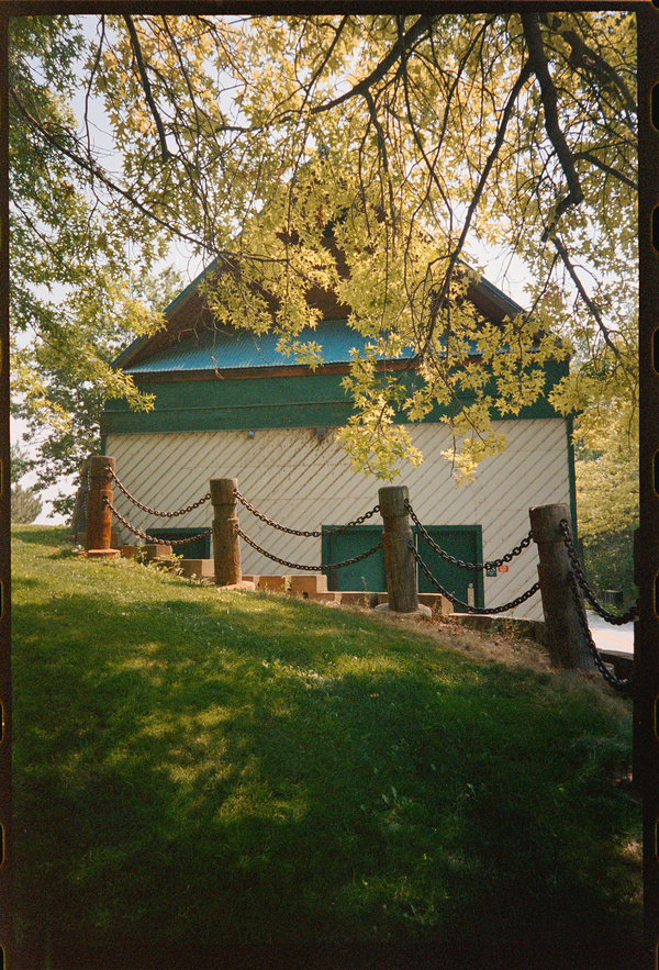 A small building with a green roof and diagonal siding is partially shaded by tree branches while surrounded by a chain-linked post fence on a grassy slope.