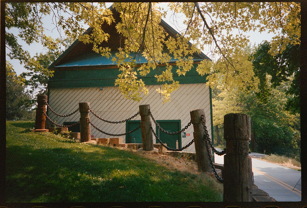 A quaint wooden building with green trim is partially obscured by leafy tree branches, while a chain-linked wooden post barrier lines a grassy slope beside a road.