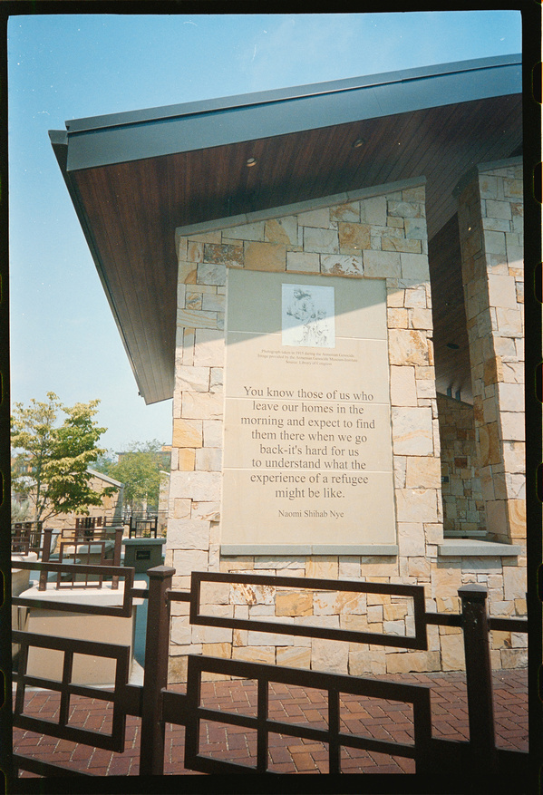 A stone building features a plaque with a quote by Naomi Shihab Nye about the challenges faced by refugees, surrounded by a metal fence and small trees.