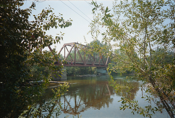 A rusted metal truss bridge spans across a calm river, surrounded by trees and greenery.
