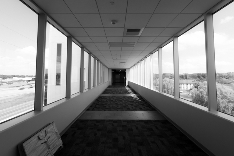 A black and white photo looking down a long, enclosed skywalk. Large windows line both sides of the hallway, showing trees and buildings outside. The perspective creates leading lines that draw the eye to an elevator at the end of the hall. A folded package delivery tote lays against the left wall.