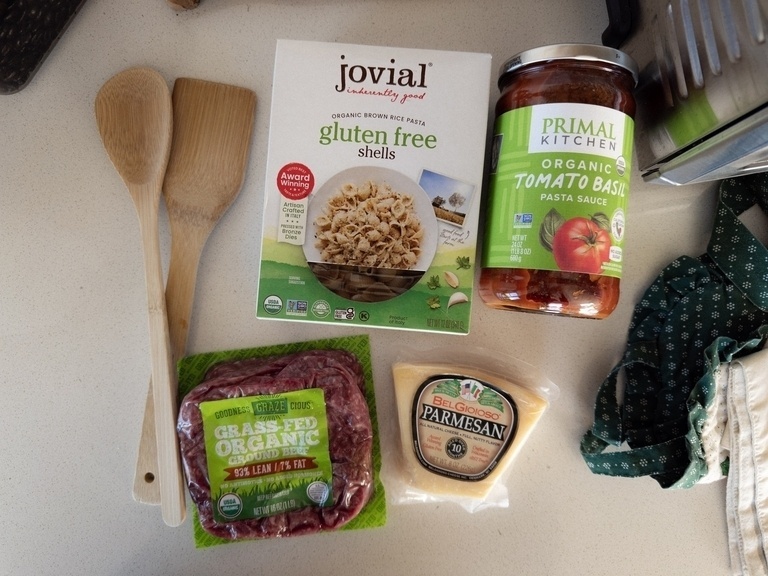 Overhead on a light countertop: two wooden cooking spoons crossed at the top left; a box of Jovial organic brown rice gluten-free pasta shells at center; a jar of Primal Kitchen organic tomato basil pasta sauce at top right; a package of grass-fed organic ground beef at bottom left; and a wedge of BelGioioso Parmesan cheese at bottom right.