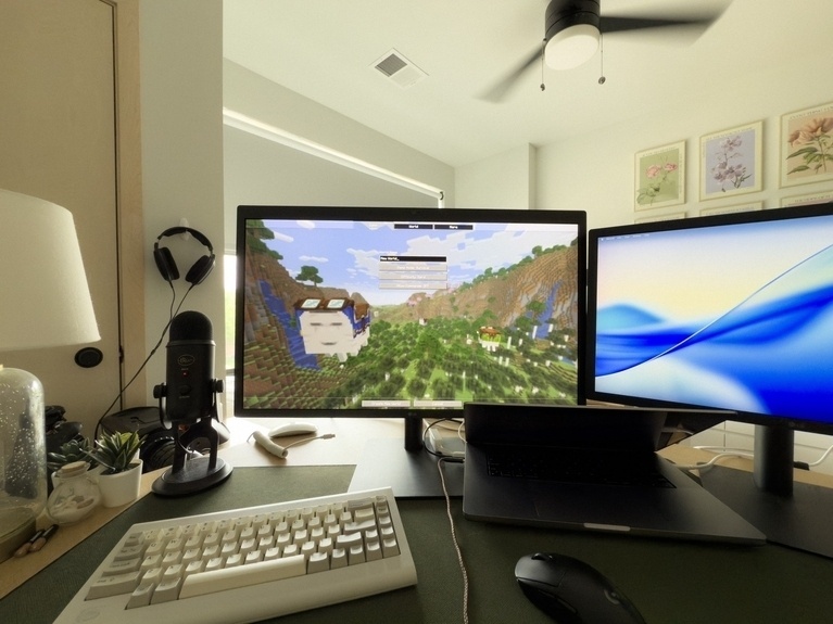 A first-person view of a desk setup shows two monitors, a keyboard, a mouse, a laptop, and a microphone. The left monitor displays the Minecraft world creation screen. The room is brightly lit, with sunlight streaming in from a large half-covered window.