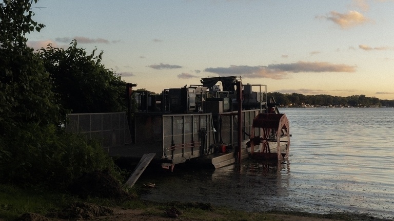 A serene lake scene during the golden hour, with the soft light of a setting sun casting a glow over the calm water. In the foreground, a mechanical harvester boat is docked on the muddy shore, its dark silhouette contrasting with the soft light, while the distant tree-lined shore is visible under a pale, dusky sky.