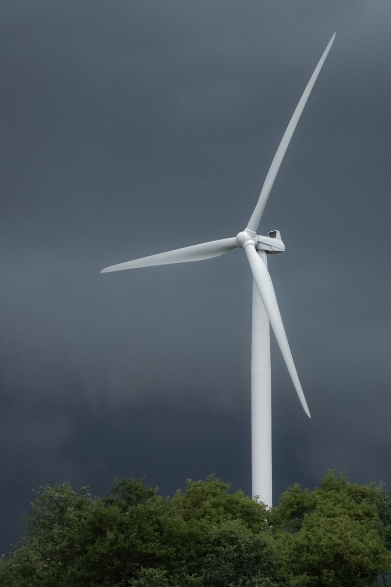 A single, large white wind turbine stands tall against a dark, stormy grey sky. The turbine's three blades are angled, suggesting it's moving slowly. The lower third of the image shows a line of vibrant green treetops, contrasting with the dramatic sky and the stark white of the turbine.